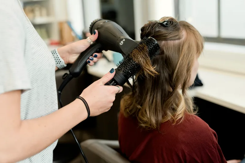 A stylist blow-drying a client's hair in a salon chair.